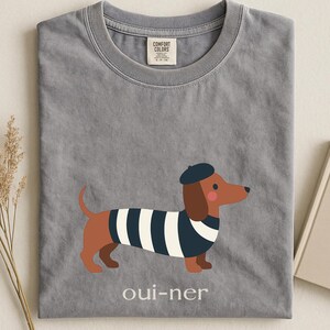 May include: Gray t-shirt with a cartoon dachshund wearing a beret and a striped shirt. The text "oui-ner" is printed below the dog. The shirt has a Comfort Colors tag.