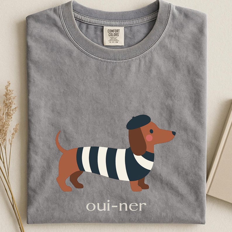 May include: Gray t-shirt with a cartoon dachshund wearing a beret and a striped shirt. The text "oui-ner" is printed below the dog. The shirt has a Comfort Colors tag.