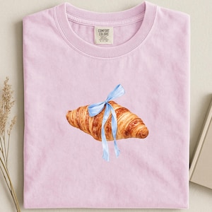 Puede incluir: Camiseta rosa claro con una ilustración en acuarela de un cruasán atado con una cinta azul claro. La camiseta tiene cuello redondo y una etiqueta que dice "Comfort Colors". La camiseta está doblada cuidadosamente.
