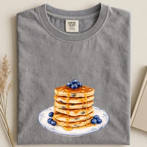 May include: Gray Comfort Colors t-shirt featuring a stack of pancakes with blueberries and syrup on a white plate with a blue floral design. The pancakes are golden brown and the blueberries are a deep blue color.