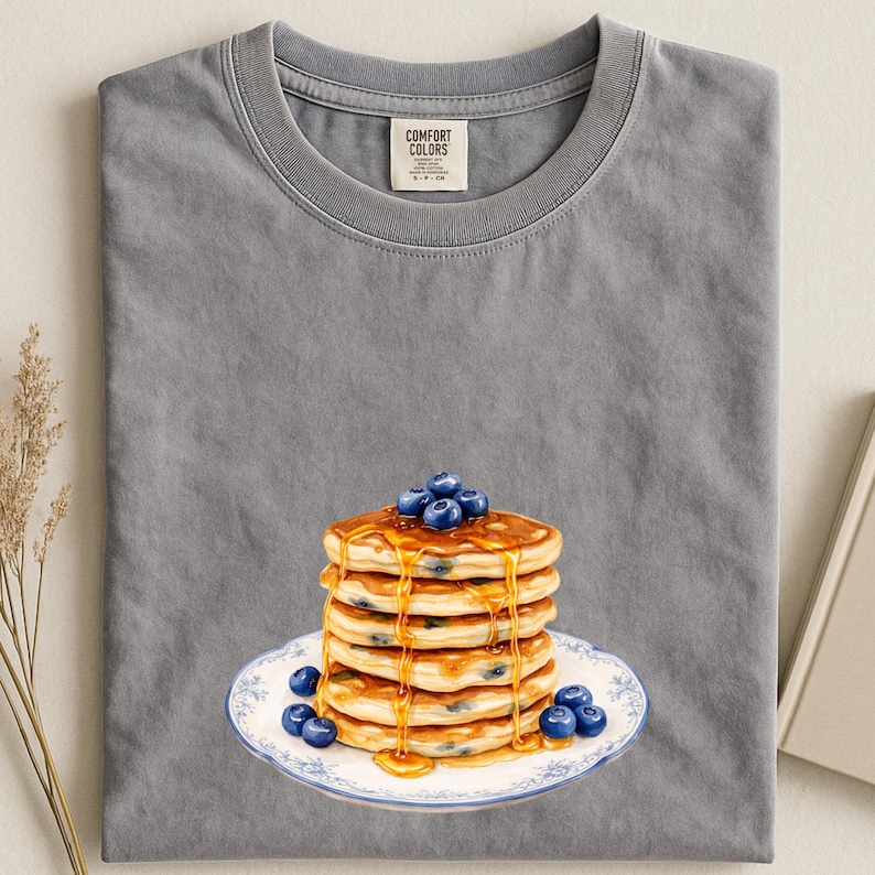 May include: Gray Comfort Colors t-shirt featuring a stack of pancakes with blueberries and syrup on a white plate with a blue floral design. The pancakes are golden brown and the blueberries are a deep blue color.