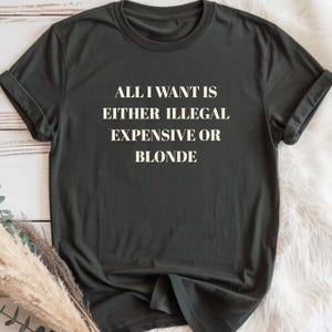 May include: Dark gray t-shirt with the text "ALL I WANT IS EITHER ILLEGAL EXPENSIVE OR BLONDE" in white block letters. The shirt has rolled-up sleeves and is displayed on a white surface.