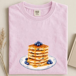 May include: A light pink t-shirt featuring a stack of pancakes with blueberries and syrup on a decorative plate. The shirt has a crew neck and a tag that reads "Comfort Colors".