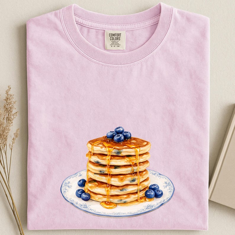 May include: A light pink t-shirt featuring a stack of pancakes with blueberries and syrup on a decorative plate. The shirt has a crew neck and a tag that reads "Comfort Colors".