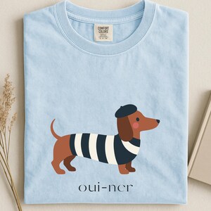 May include: Light blue t-shirt featuring a cartoon dachshund wearing a beret and a striped shirt. The dachshund has brown fur and pink cheeks. The text "oui-ner" is printed below the dog.