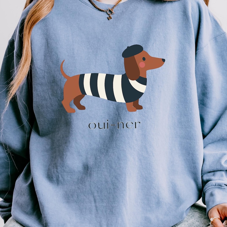 May include: A light blue sweatshirt featuring a cartoon dachshund wearing a black beret and a black and white striped shirt. The text "oui-ner" is printed below the dog.