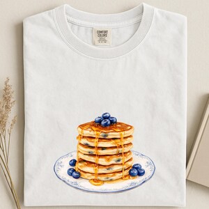 May include: White t-shirt featuring a watercolor illustration of a stack of pancakes with blueberries and syrup. The pancakes are on a plate with a blue floral design. The shirt has a Comfort Colors tag.