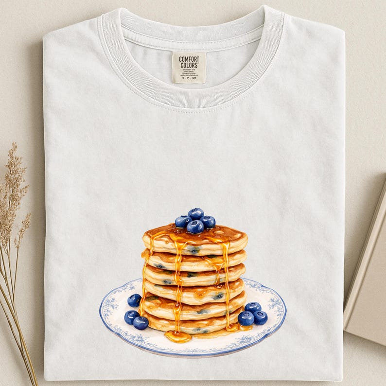 May include: White t-shirt featuring a watercolor illustration of a stack of pancakes with blueberries and syrup. The pancakes are on a plate with a blue floral design. The shirt has a Comfort Colors tag.