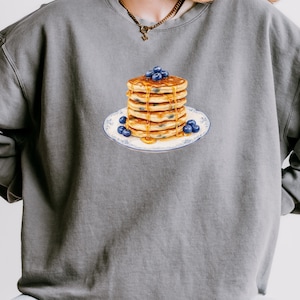 May include: A gray sweatshirt featuring a stack of pancakes with blueberries and syrup on a decorative plate. The pancakes are golden brown, and the blueberries are a deep blue. The plate has a blue floral design.
