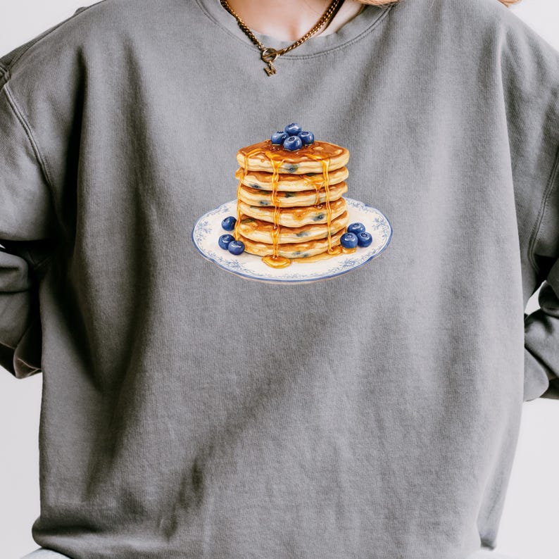 May include: A gray sweatshirt featuring a stack of pancakes with blueberries and syrup on a decorative plate. The pancakes are golden brown, and the blueberries are a deep blue. The plate has a blue floral design.