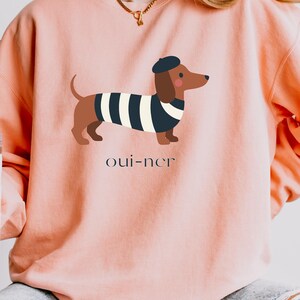 May include: Pink sweatshirt featuring a cartoon dachshund wearing a beret and a striped shirt. The dog illustration is brown, black, and white. The text "oui-ner" is printed below the dog.
