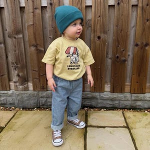 May include: A child in a teal beanie, yellow t-shirt with a "Lemonade Stroller" graphic, and blue striped trousers. The child wears brown and white trainers. The background features a wooden fence.