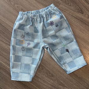 May include: Light blue trousers with an elasticated waistband and a checkered pattern. The fabric features small embroidered Halloween-themed icons including a ghost, a skull, a haunted house, a spider web, a spider, a lollipop, and a skeleton hand.