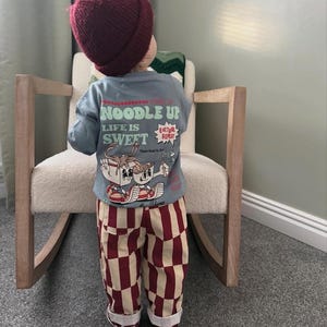 May include: A child in a maroon beanie, a light blue long-sleeve shirt with the text "NOODLE UP LIFE IS SWEET" and a cartoon graphic, and red and cream checkered trousers. The child is standing in front of a cream rocking chair.