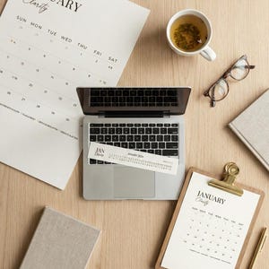 May include: A workspace scene featuring a laptop, calendar, clipboard, and a cup of tea. The calendar and clipboard display "January Clarity". There are also glasses and a pen on the wooden surface.