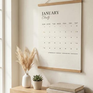 May include: A minimalist wall calendar for January, with the word "Clarity" in cursive. The calendar is suspended by a wooden rod and a string. A wooden shelf below holds a vase with dried flowers, a small potted succulent, and two books.