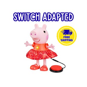 May include: A pink Peppa Pig character toy wearing an orange top with a yellow flower and a red skirt with white star patterns. The toy is switch adapted with a black cord connected to a round red button. The text SWITCH ADAPTED is visible.