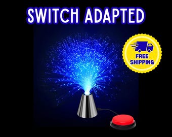 Op de afbeelding: Een schakelaar-aangepaste glasvezellamp met een zilverkleurige conische voet en een felblauwe lichtweergave. Het apparaat is verbonden via een zwart snoer met een ronde rode drukknopschakelaar. De tekst "SWITCH ADAPTED" staat bovenaan in het wit.