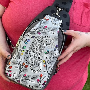 May include: A gray sling bag with a black polka dot strap and a zipper closure. The bag features a detailed print of a leopard amidst floral elements in gray, white, and colorful accents. The bag is held by a person wearing a red shirt.