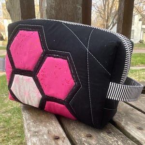 May include: A black and pink quilted bag with a geometric design. The bag features a hexagon pattern in bright pink and white, outlined in black. It has a black and white striped handle and zipper.