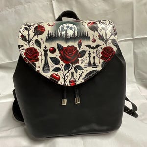 May include: Black backpack with a flap featuring a floral and gothic design. The flap has red roses, chess pieces, and a full moon. The bag has a drawstring closure and adjustable straps.