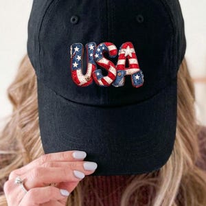May include: Black baseball cap with the word "USA" embroidered on the front. The letters are designed with red, white, and blue patterns, resembling the American flag. The cap has a curved brim and a classic, casual style.