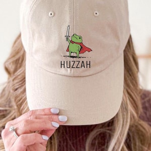 May include: A tan baseball cap featuring a cartoon frog with a sword and cape. The frog illustration is green with a red cape. The word "Huzzah" is printed below the frog. The cap is made of a soft material.