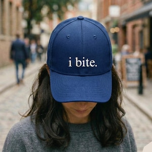 May include: A navy blue baseball cap with the text "i bite." in white. The cap has a curved brim and a structured fit. The hat is a casual accessory.