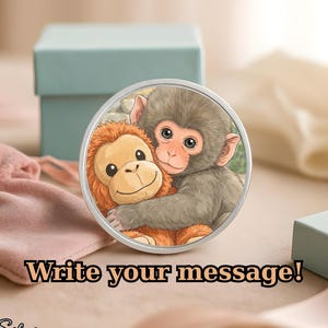 May include: A round silver pendant featuring a cartoon illustration of two monkeys hugging. The pendant has the text "Write your message!" and "Silver" in a script font. A light blue gift box and a pink pouch are in the background.