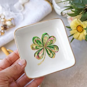 May include: A square, white ceramic trinket dish with gold trim. The dish features a colorful shamrock design with green, pink, and gold accents. The dish is held in a hand, with a blurred background including flowers and a white towel.