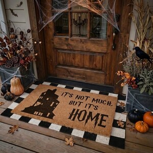 May include: A rectangular coir doormat with a black silhouette of a multi-story house and the text "IT'S NOT MUCH, BUT IT'S HOME" in a stylized font. The mat is layered over a black and white checkered rug in front of a wooden door.