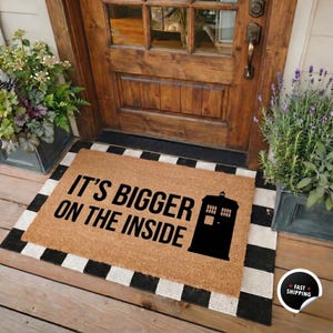 May include: A rectangular tan coir doormat featuring the black text IT'S BIGGER ON THE INSIDE and a black silhouette of a police box. The mat is layered over a black and white checkered rug, positioned in front of a wooden door with glass panels.