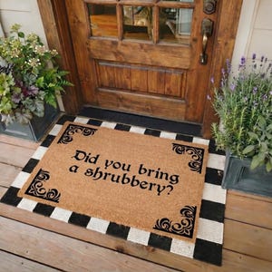 May include: A rectangular tan coir doormat with black decorative corner flourishes and the black text Did you bring a shrubbery? The mat is layered over a black and white checkered rug in front of a wooden door with a metal handle and glass panels.