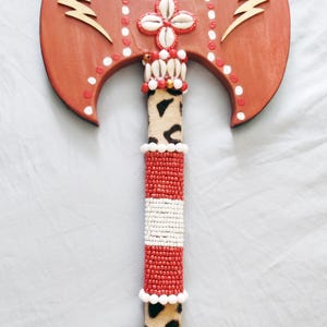 May include: A decorative axe with a reddish-brown blade and handle. The blade features gold lightning bolt designs and white dot accents. The handle is adorned with red and white beads, animal print fabric, and shell embellishments.