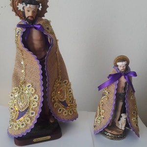 May include: Two statues of San Lazaro. The larger statue has a brown burlap cape with gold sequins and purple trim. The smaller statue has a similar cape and a small dog at its feet. Both statues have a gold halo.