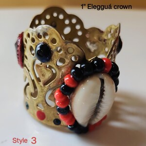 May include: A gold-colored metal crown, approximately 1 inch in diameter, with a decorative openwork design. The crown is adorned with black and red beads, a white seashell, and black and red accents. The text "1" Elegguá crown" and "Style 3" are visible.