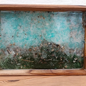 May include: A rectangular wooden tray with a turquoise and green resin design. The resin incorporates small stones, twigs, and other natural elements. The tray has a natural wood frame and integrated handles.