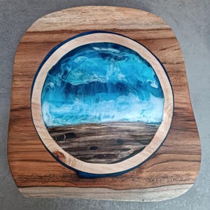 Könnte beinhalten: Holzservierbrett mit kreisförmigem Ozean-Harz-Design. Das Brett hat einen natürlichen Holzmaserungsrand und ein zentrales Epoxidharz-Design, das Meereswellen in Blau- und Weißtönen darstellt. Ein einzigartiges Dekorationsstück.