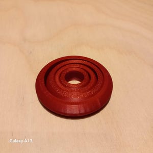 Könnte beinhalten: Ein rotes, 3D-gedrucktes Objekt mit einer Reihe konzentrischer Ringe. Das Objekt ist kreisförmig und hat eine strukturierte Oberfläche. Das Design weist ein zentrales Loch auf und ist wahrscheinlich ein dekorativer oder funktionaler Gegenstand.