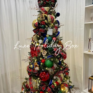 May include: A decorated Christmas tree featuring red, green, and gold ornaments, plaid ribbon, and small teddy bear accents. The tree is illuminated with warm lights and topped with a festive spray. The text Luxe Holiday Box is overlaid in white.