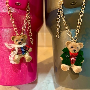 May include: Two gold chain charms with teddy bear pendants. One bear wears a plaid sweater and scarf, the other a green jacket with "RL" on it. The charms are attached to a pink and a gray tumbler.
