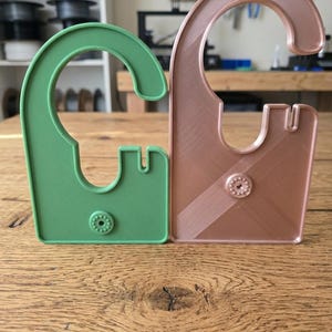 May include: Two C-shaped plastic hangers, one in matte green and one in metallic copper. Each hanger features a curved top hook, a central cutout, a small notched side section, and a circular textured button detail at the base of the flat body.