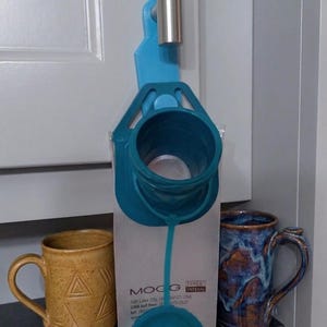 May include: A teal plastic enteral feeding bag holder hangs from a silver metal hook. The bag features a teal screw-top opening, a teal cap, and printed text reading MOOG, TEYEX ENTERAL, Salt Lake City, UT 84123 USA, and a toll-free phone number.