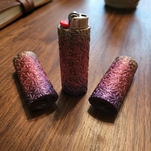 May include: Three cylindrical lighter cases featuring a textured, embossed leaf pattern. The cases display a gradient color transition from a deep purple at the base to a vibrant red and gold finish at the top. One case is upright with a metal lighter insert.