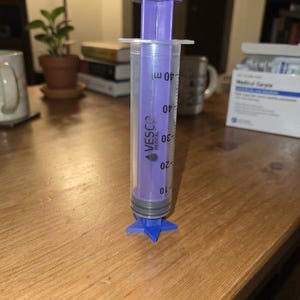 Peut inclure: Une seringue orale en plastique transparent avec un piston violet et une base en forme d'étoile. Le corps présente des graduations noires jusqu'à 40 ml et le texte VESCO MEDICAL imprimé verticalement sur la tige du piston violet.