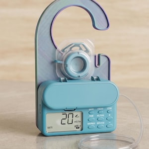 May include: A light blue enteral feeding pump with a digital display showing 20 ml/hr, a control panel with buttons labeled ON/OFF, FEED INT, CLEAR, PRIME, VOL/TOTAL, RUN/PAUSE, and RATE/DOSE, attached to a large, iridescent, hook-shaped hanger.