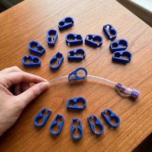 May include: A collection of small, blue, 3D-printed plastic clips with a figure-eight shape, designed to hold thin, clear tubing. One clip is shown attached to a piece of flexible, transparent plastic tubing that features a purple connector at the end.