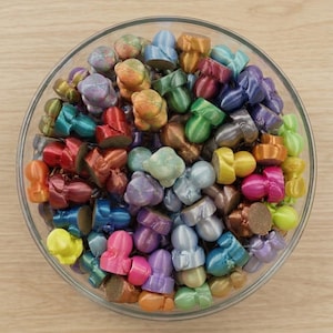 May include: A clear glass bowl filled with a large collection of small, mushroom-shaped objects in a variety of metallic and solid colors, including blue, green, pink, yellow, orange, purple, and bronze, each with a flat, textured base.