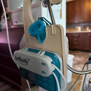 May include: A beige plastic hanger support for an Infinity Moog enteral feeding pump. The device features a teal and white housing with the text Infinity Moog, a teal cap, and a clear fluid bag attached to the bottom, suspended from a metal pole.