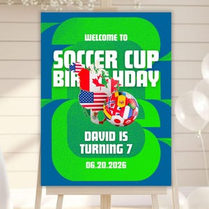 以下が含まれることがあります： イーゼルに飾られたサッカーをテーマにした誕生日パーティーのサイン。サインは青と緑で、「Welcome to Soccer Cup Birthday」と「David is turning 7」という文字が書かれています。サッカーボールと旗も描かれています。